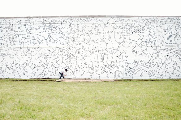 Thin black lines trace a web of simplified faces and stick figures connected by abstract, looping patterns against a white-painted brick wall. The wall fills the top half of the image, and an expanse of light green grass fills the bottom half. The artist, a black woman wearing a white shirt and black jeans, appears bent over with spray can in hand slightly to the left of center; she is dwarfed by the scale of the wall.