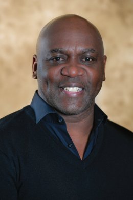 Photograph of a smiling black man wearing a black collard shirt that is slightly open at the neck