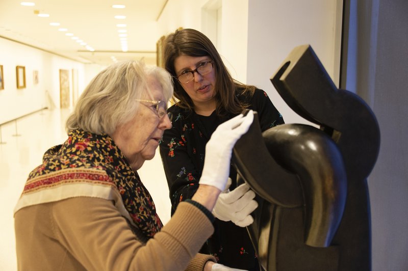 An older woman with white gloves touches a large black sculpture while a younger woman with glasses looks on