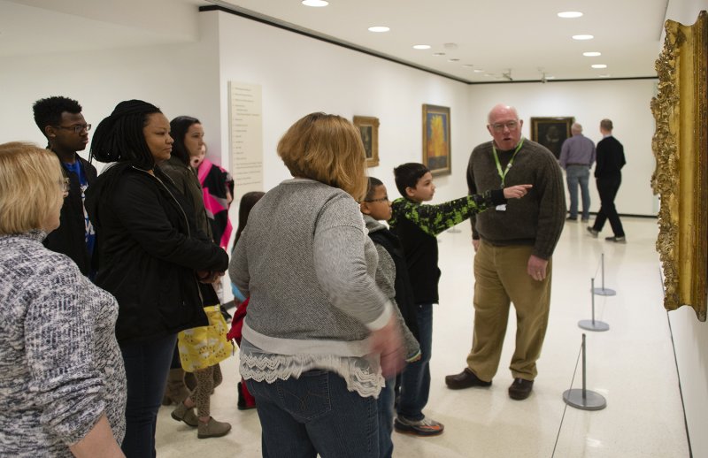 People of all different ages on a tour. One boy points toward the painting they are all looking at.