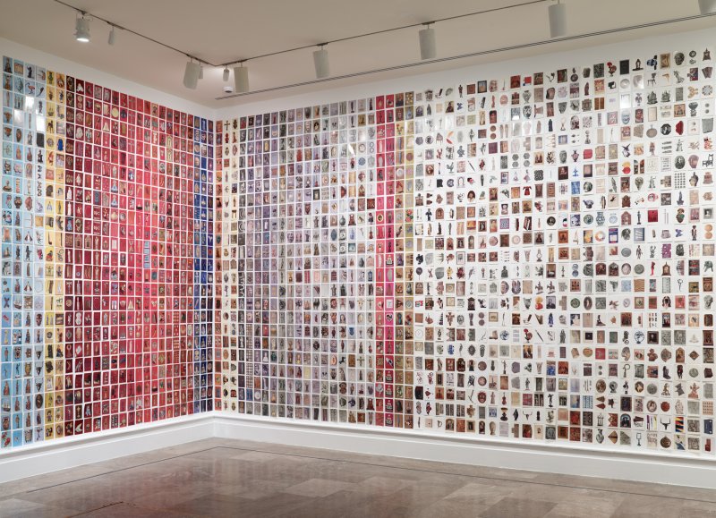 A corner of a room with postcards of museum objects on different colored backgrounds fixed to the walls, from floor to ceiling