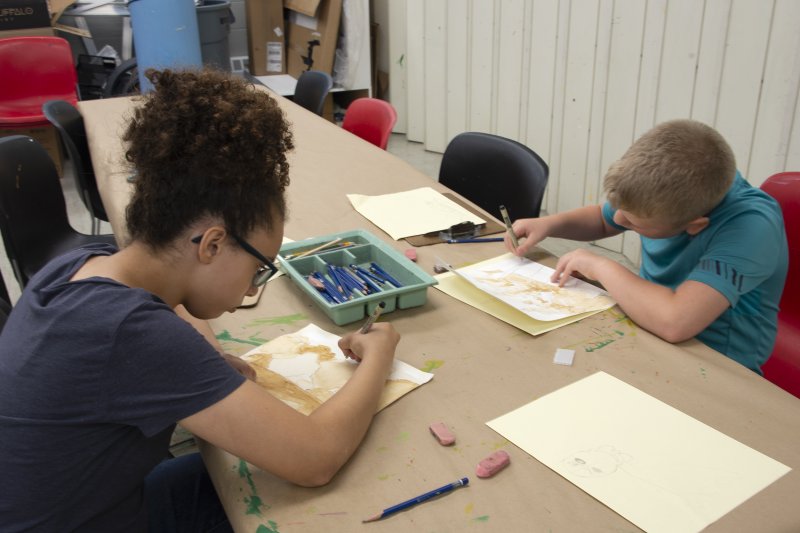 A young teen girl and a young teen boy drawing at a long table