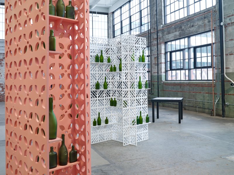 Two wooden screens, painted pink and white, holding green glass bottles, with a small square table in the background