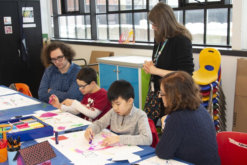 Two children sit at a table making art while two adults sit next to them and one adult stands behind them to help guide them