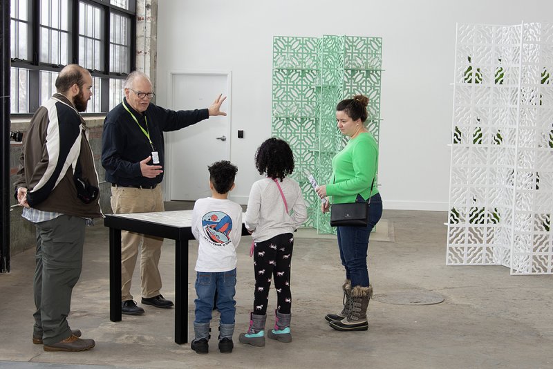 Two men, a woman, and two kids looking at two wooden screens with glass bottles placed on shelves