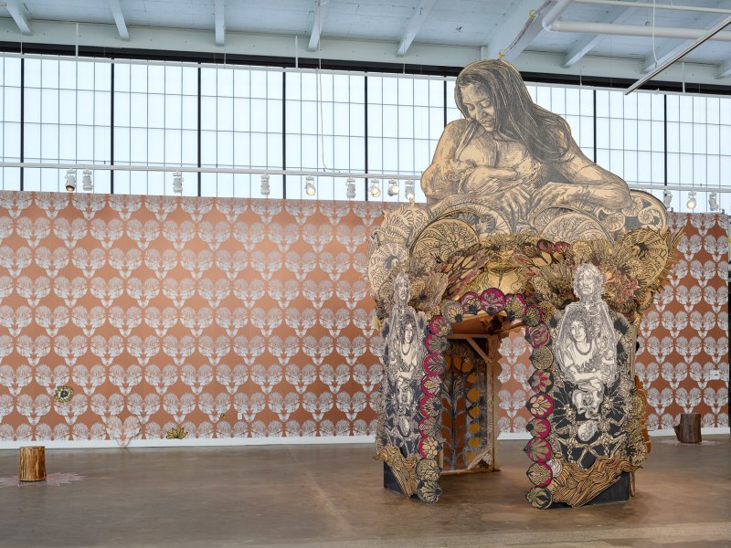 A photograph of a temple-like structure with a sculpture of a woman nursing a baby on top. In the background is bronze and white wallpaper in a repeating pattern.
