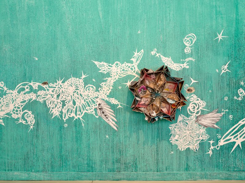 A segmented wheel affixed to a teal wall with intricate white paper cutouts surrounding it