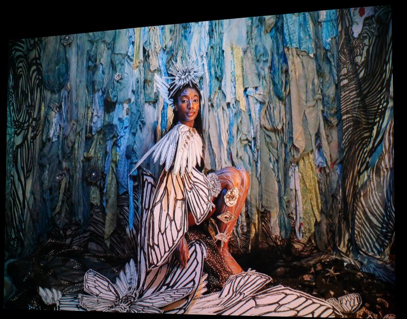 A Black woman sitting sideways with her head turned to look at the camera. She has white paper wings with black outlines attached to her arms.