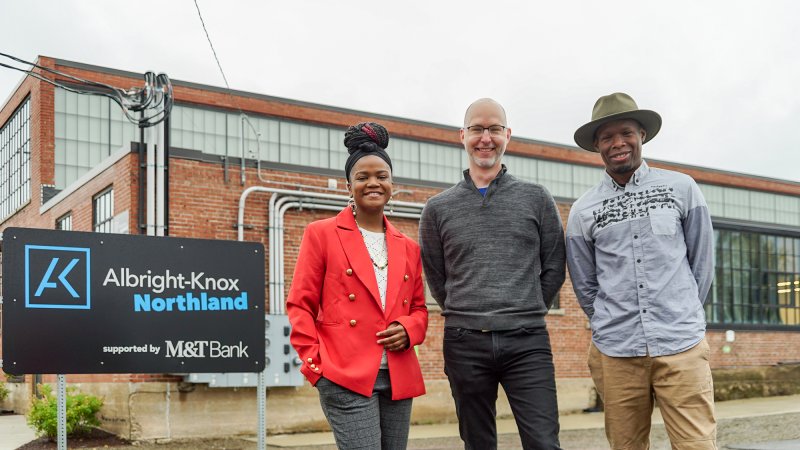 A Black woman in a red jacket, a white man in a gray sweater, and a Black man in a blue shirt and hat standing in front of a large brick building