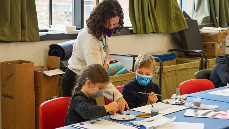 A teacher helping two kids paint different shades at a table