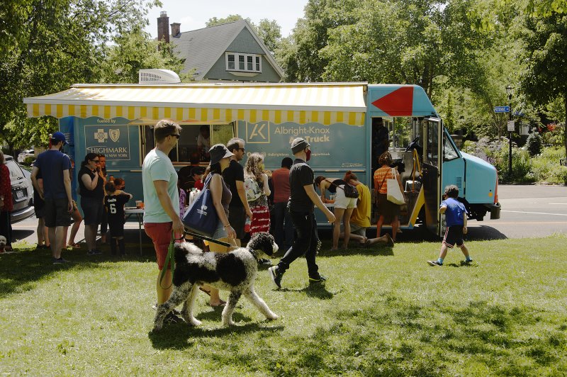 People standing and walking in front of a large blue truck parked on a road next to a big grassy area