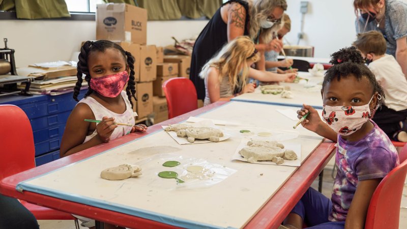Two girls making sculptures at a table, looking up to smile for the camera
