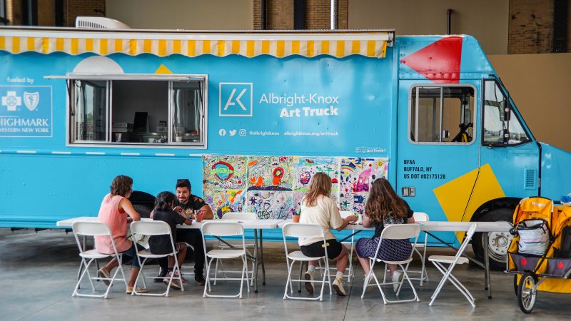 Adults and kids making art a long white table in front of a big blue truck