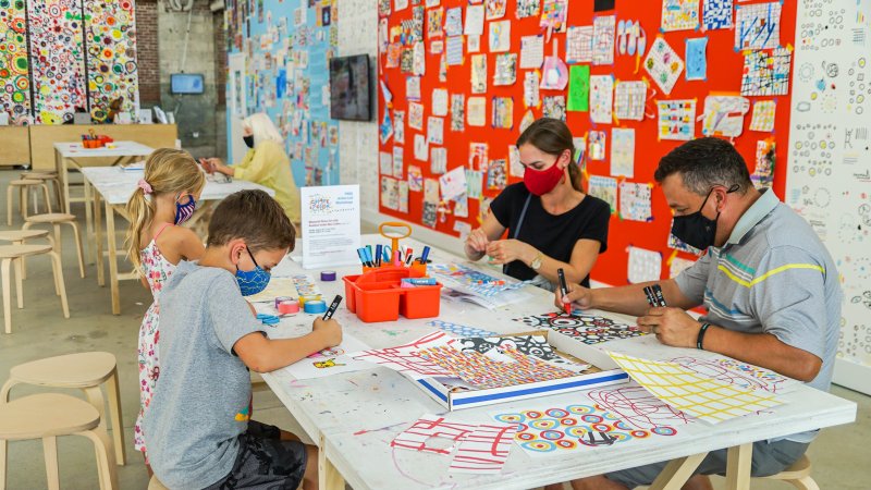 A family making art at a big table, with art hanging on a red wall behind them