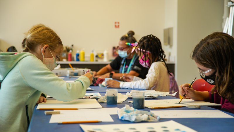 Three girls and one woman painting at a long table, with face masks on