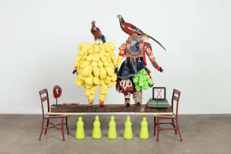 Two sculpted figures made from mixed materials stand on a board suspended between two chairs, below a row of matriarchal statuettes, perched on the figures shoulders are two bird statues, possible peacocks