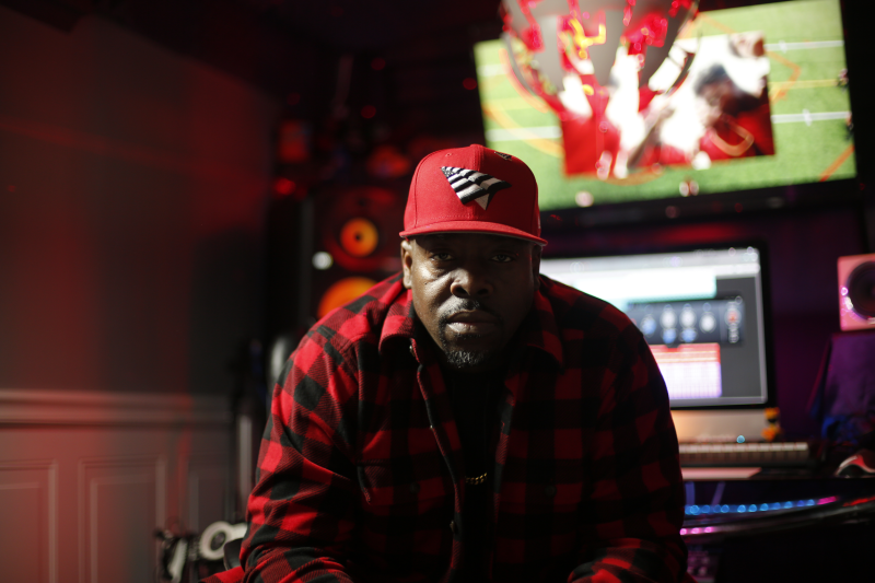 A man of dark skin tone and goatee in Buffalo plaid shirt and red hat is seated, shown from the waste up, he is lit from behind by out of focus monitors