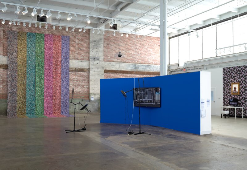 Installation view of three artworks: one with seven colorful banners hanging from the ceiling, one with a tv screen on a wall and two pieces of AV equipment in front of it, and one with two computers on desks in front of a wall with flower patterned wallpaper and two paintings of Black men