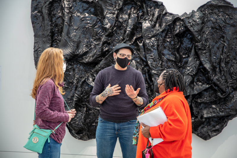 Three people standing in front of a large black sculptural work on the wall