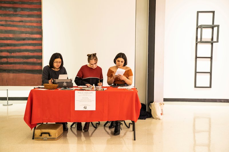Three young women sit a red desk, two writing, one typing at typewriter, in the middle of a gallery space
