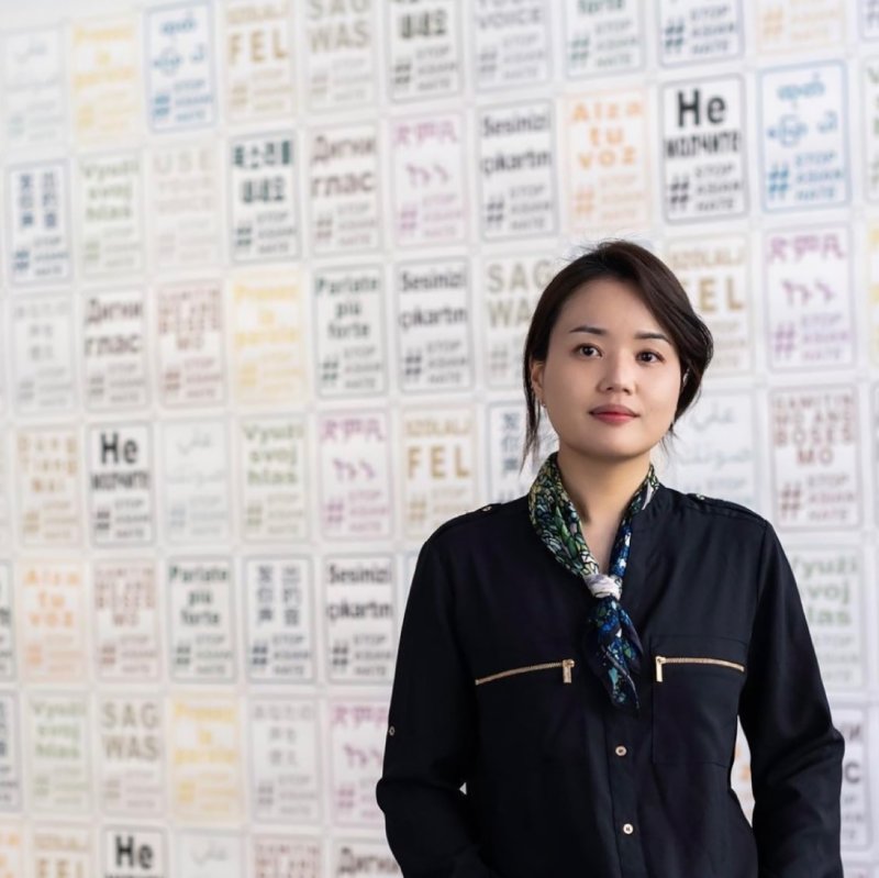 Mizin Shin, a Korean woman, stands in jacket in front of wall of her prints in multiple colors of "Use Your Voice #StopAsianHate"
