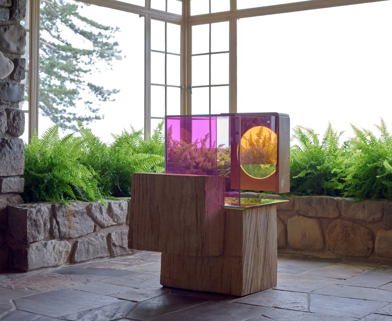 A sculpture with a wooden base and side with pink and orange plexiglass forming a box-like shape on top situated in a room in a house with stone floors and large windows