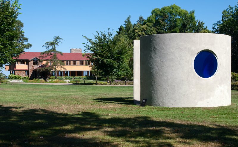 A cylindrical concrete structure with circular blue glass set in an aperture appears on the sweeping green landscape with a distinct house in the background