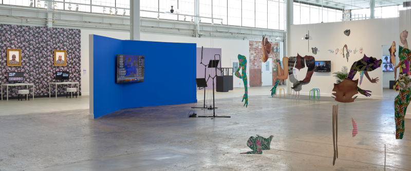 Installation view of three artworks: one with seven colorful banners hanging from the ceiling, one with a tv screen on a wall and two pieces of AV equipment in front of it, and one with two computers on desks in front of a wall with flower patterned wallpaper and two paintings of Black men