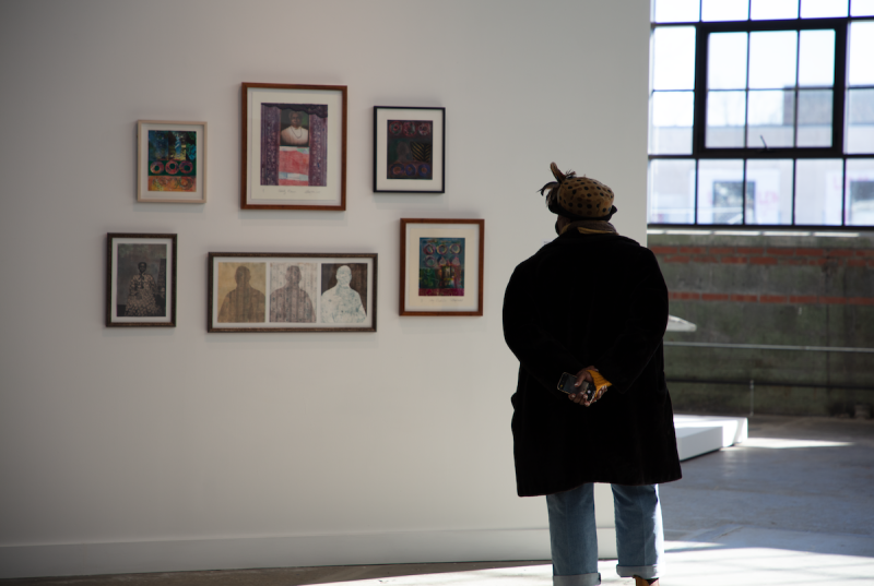 A person in a black coat stands with hands clasped behind their back looking at an array of images hung in front of them.
