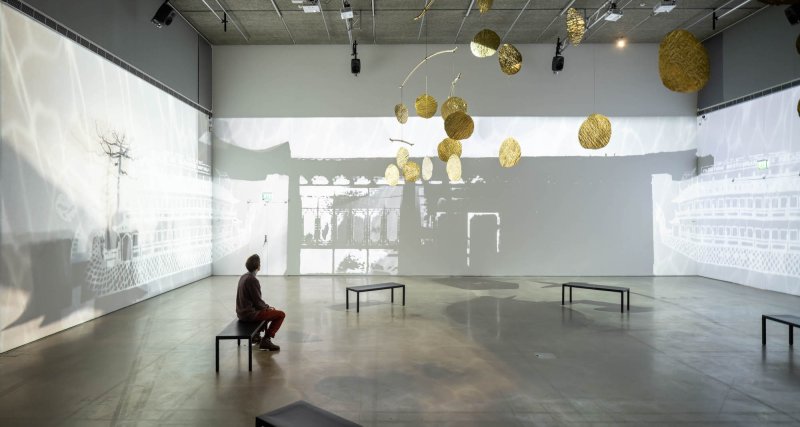 A person sits on a bench in the middle of room with black and white projections on three walls surrounding and a gold medallions hanging from the ceiling