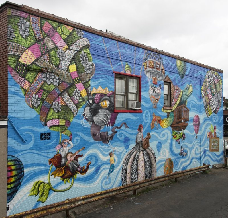 Whimsical mural on the side of a brick building featuring a blue swirly sky and multiple hot air balloons made of plants and floral patterns with people and animals riding within