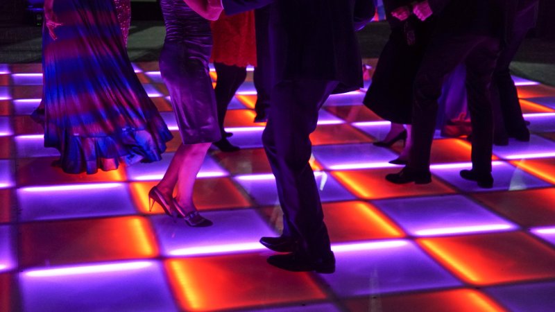 Close up of A neon orange and purple checkered dance floor with various pairs of legs dancing 
