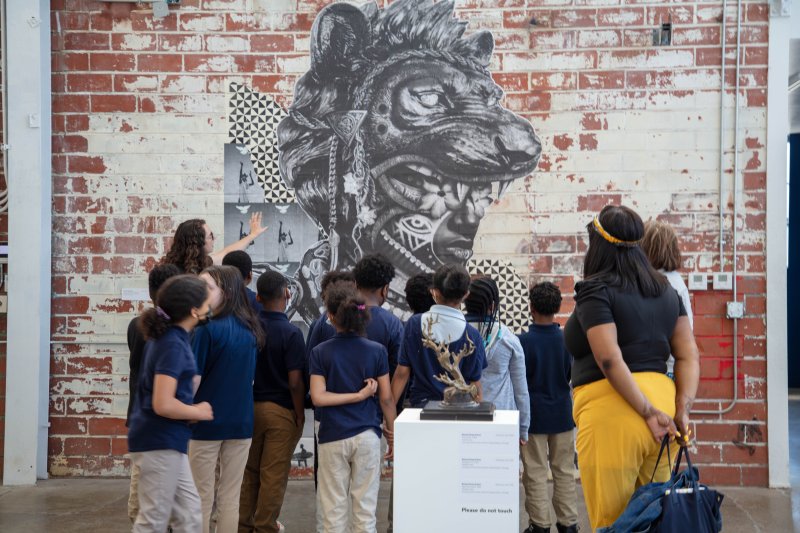 A group of students and three adults look at a black and white mural of a person's face wearing a lion mask on a brick wall
