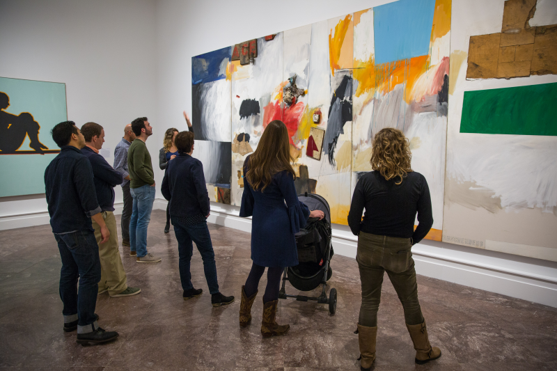 A group of eight people (including a woman with a baby stroller) admire a large abstract artwork 