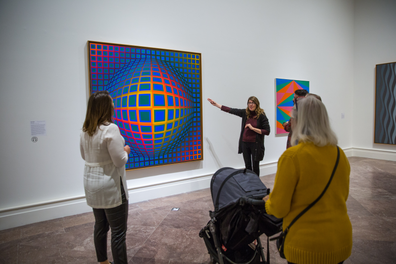 A group of four onlookers (one with a baby stroller) look at a tour guide explaining a vibrant geometric painting 