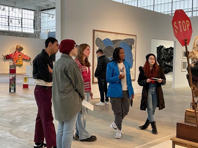 A group of five people stand around a sculpture and discuss while in a gallery
