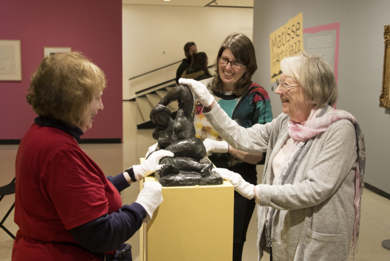 Three white women wearing gloves are smiling and touching a sculpture of a woman on a yellow pedestal