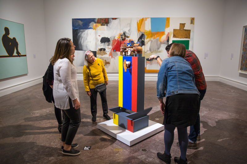 A group of four women and a man stand around a sculpture painted yellow blue red and black with wooden heads
