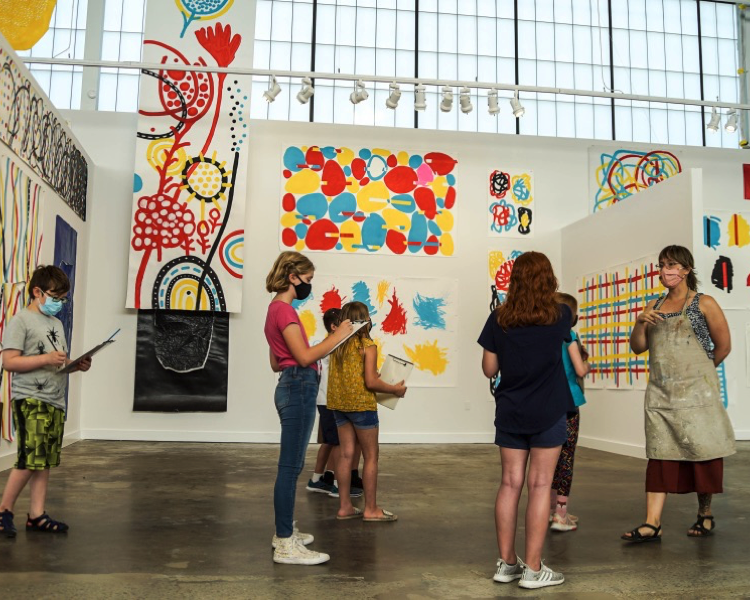 Children with clipboard and pens work while standing in a gallery with colorful artwork while listening to a educator in an apron speak