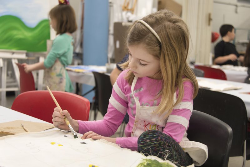 A young blonde girl in a pink shirt paints at a table