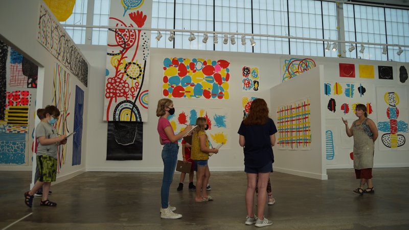 A group of young students stand and listen to an instructor with sketchpads in a colorful gallery