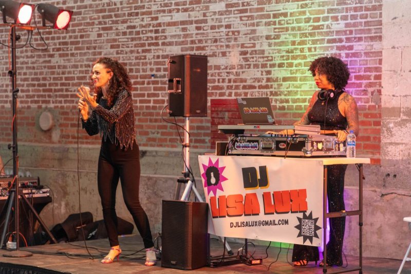 A white woman is holding a microphone on stage while an African American woman DJs behind her on stage. 