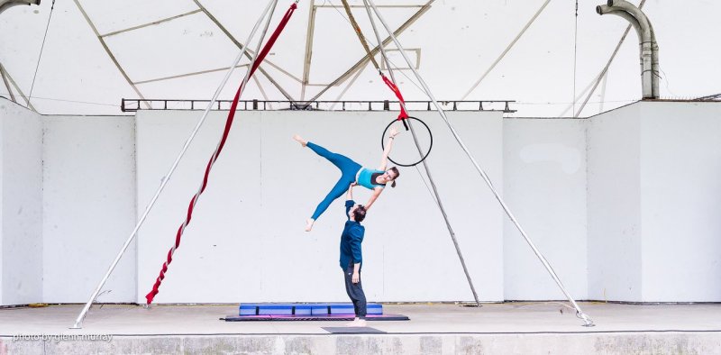 A man holding a woman up with one hand as she is reaching for an aerialist's hoop. 
