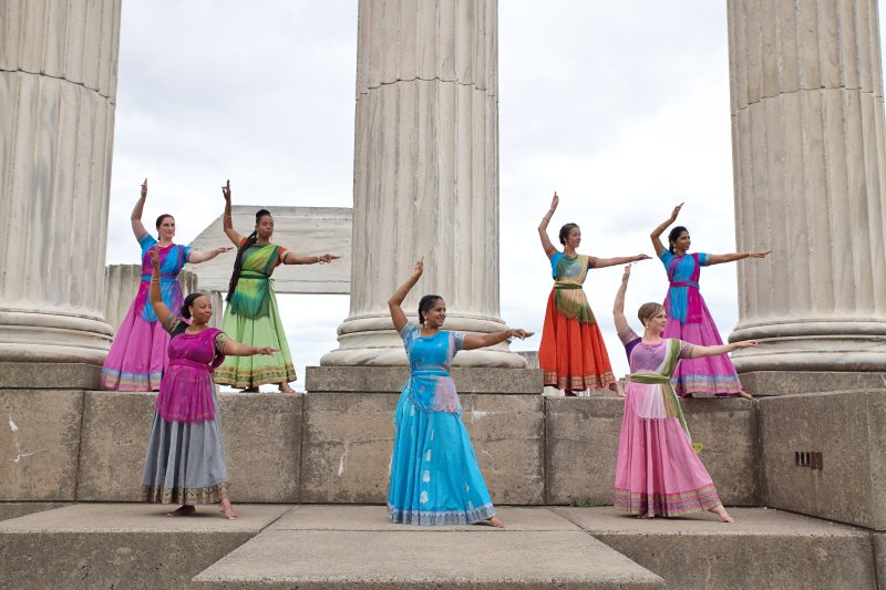 Six women in traditional saris pose in front of large marble columns 