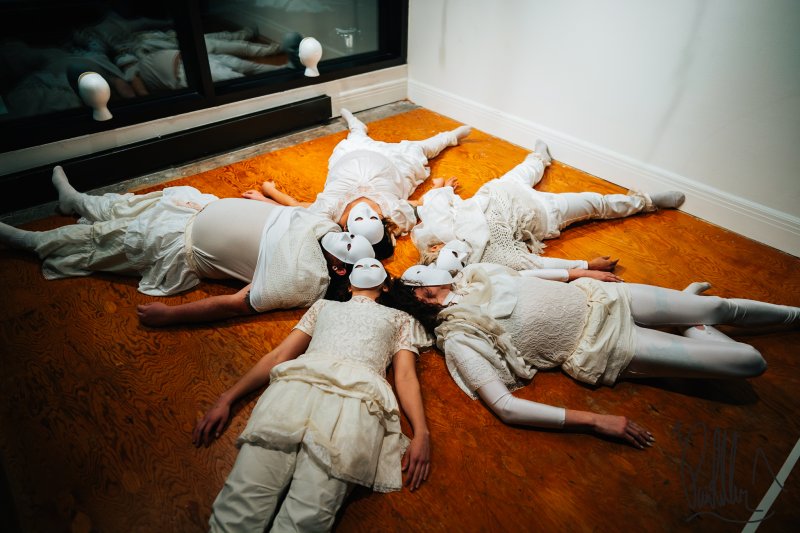 Five people in all white wearing white masks lay on the ground in a circle
