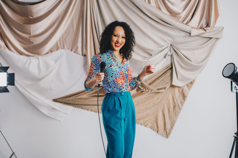 A woman with brown curly hair, wearing a patterned blue blouse and blue pants poses with a microphone