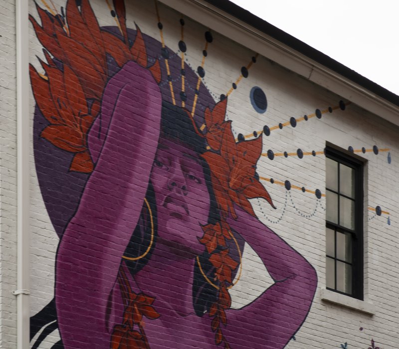 A close up of a painted mural on the side of a white brick building of a woman with purple skin and a halo design of orange