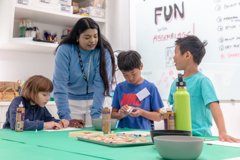 Two young boys and a girl looking towards a woman in a blue sweater leading an art project at a green table