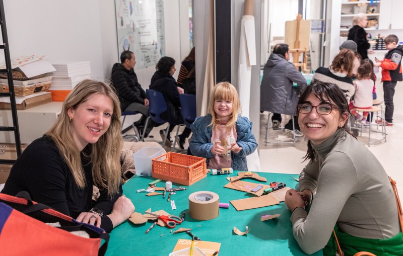 a woman with blonde hair, a woman with dark hair, and a young girl with blonde hair all smiling while sitting at a green table with art supplies