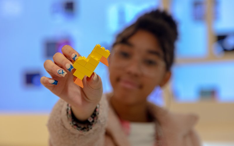 A photo of a girl (blurry) with dark curly hair in a bun holding a duck made out of yellow legos to the camera 